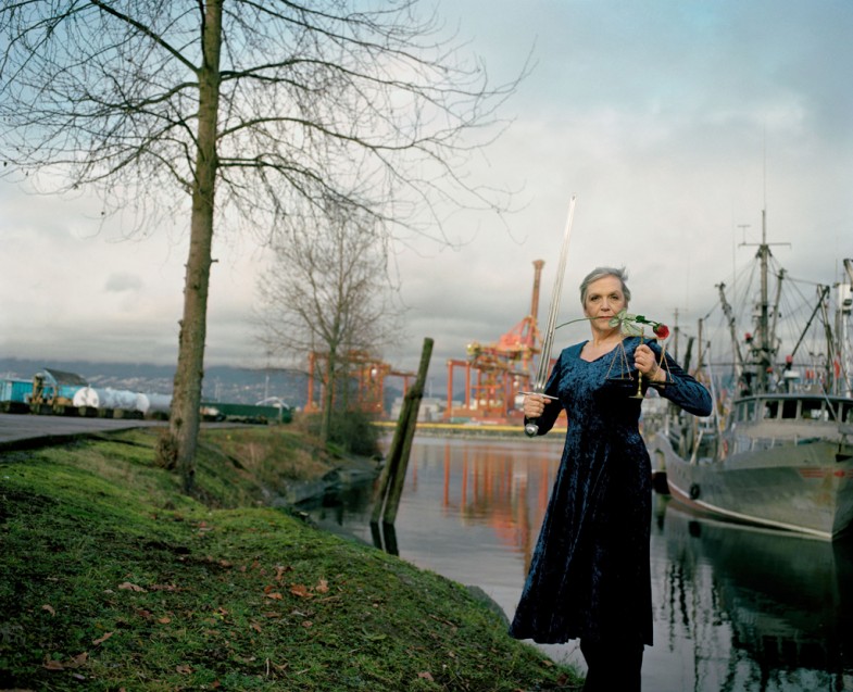 The artist with a red rose in her mouth, holding a sword and scale in the background we see boats, cranes and a cloudy sky reflected on water.